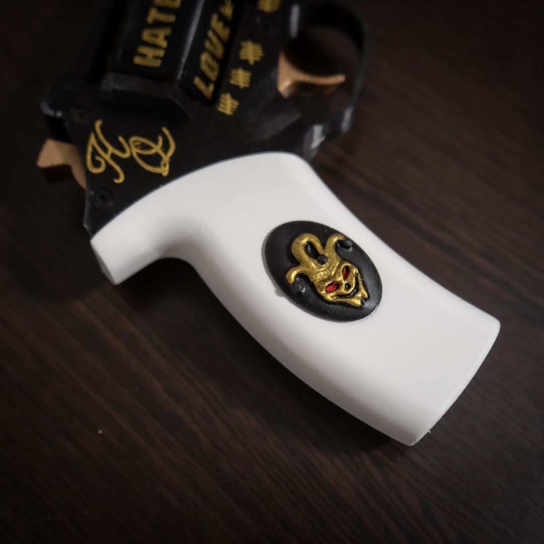 Close-up of Harley Quinn's Revolver handle with white grip and gold Joker face emblem on dark wooden surface