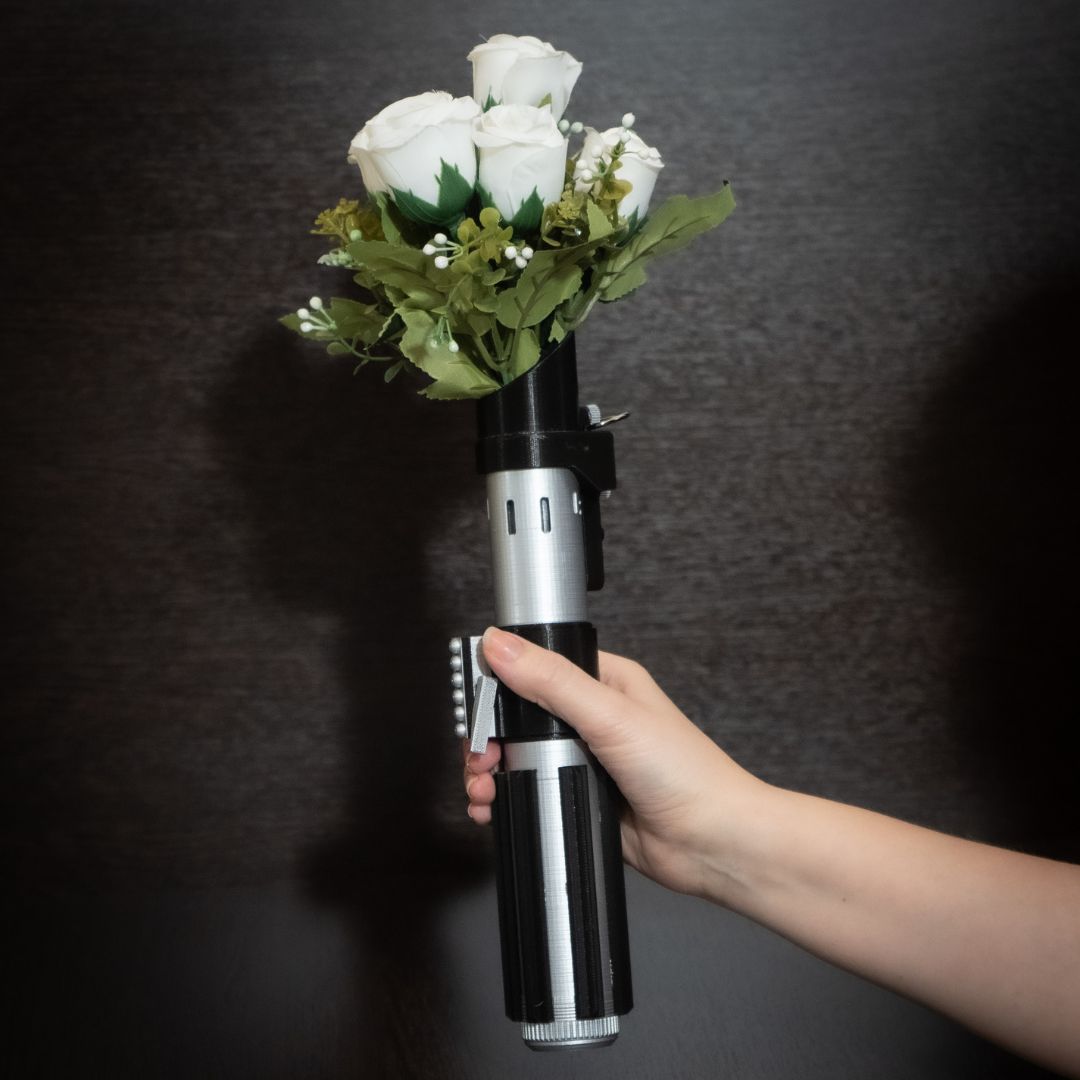 Hand holding a Star Wars inspired Darth Vader lightsaber bridal bouquet holder with white roses and greenery against a dark background