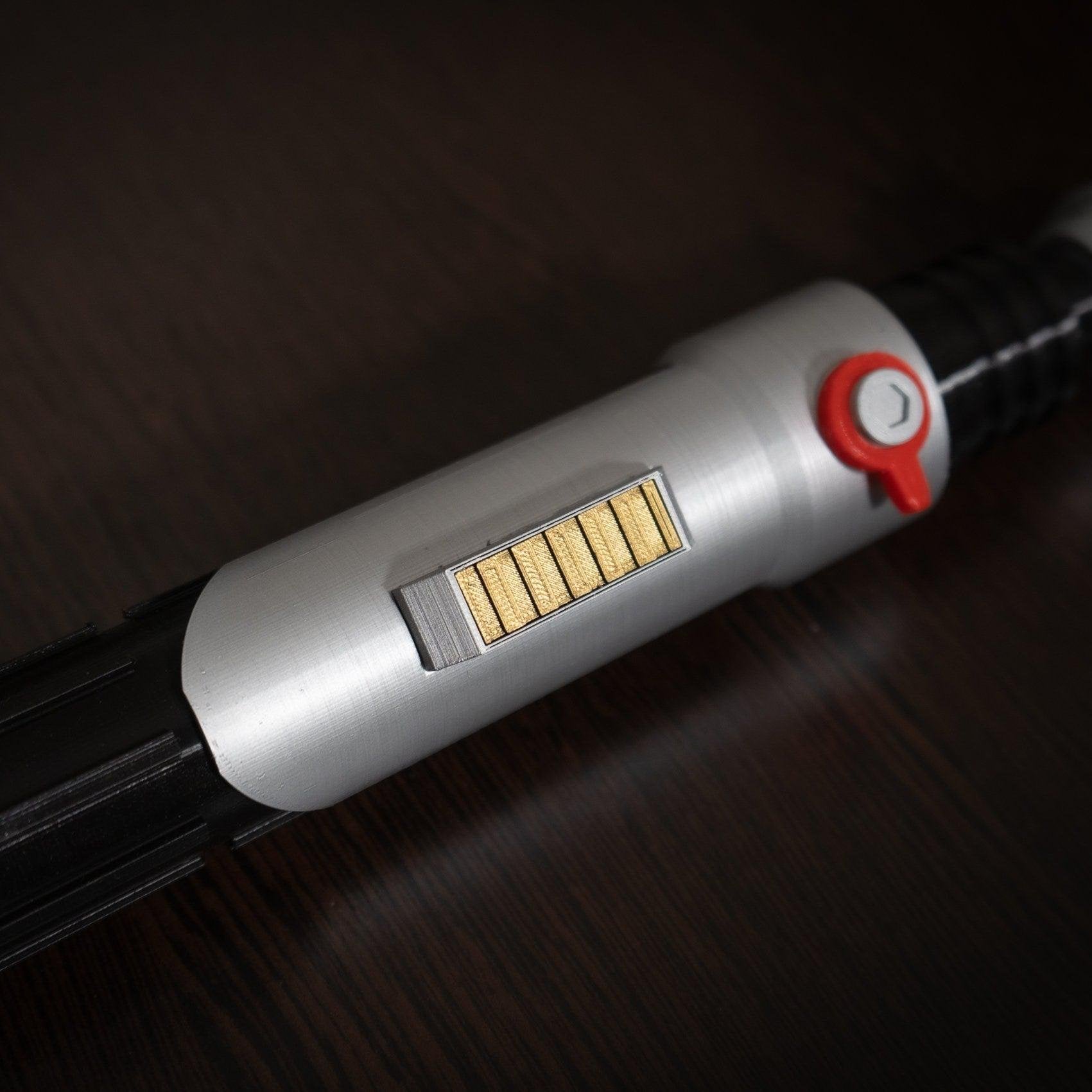 Close-up of Ezra Bridger second lightsaber hilt cosplay prop with silver body, gold details, and red button on dark surface