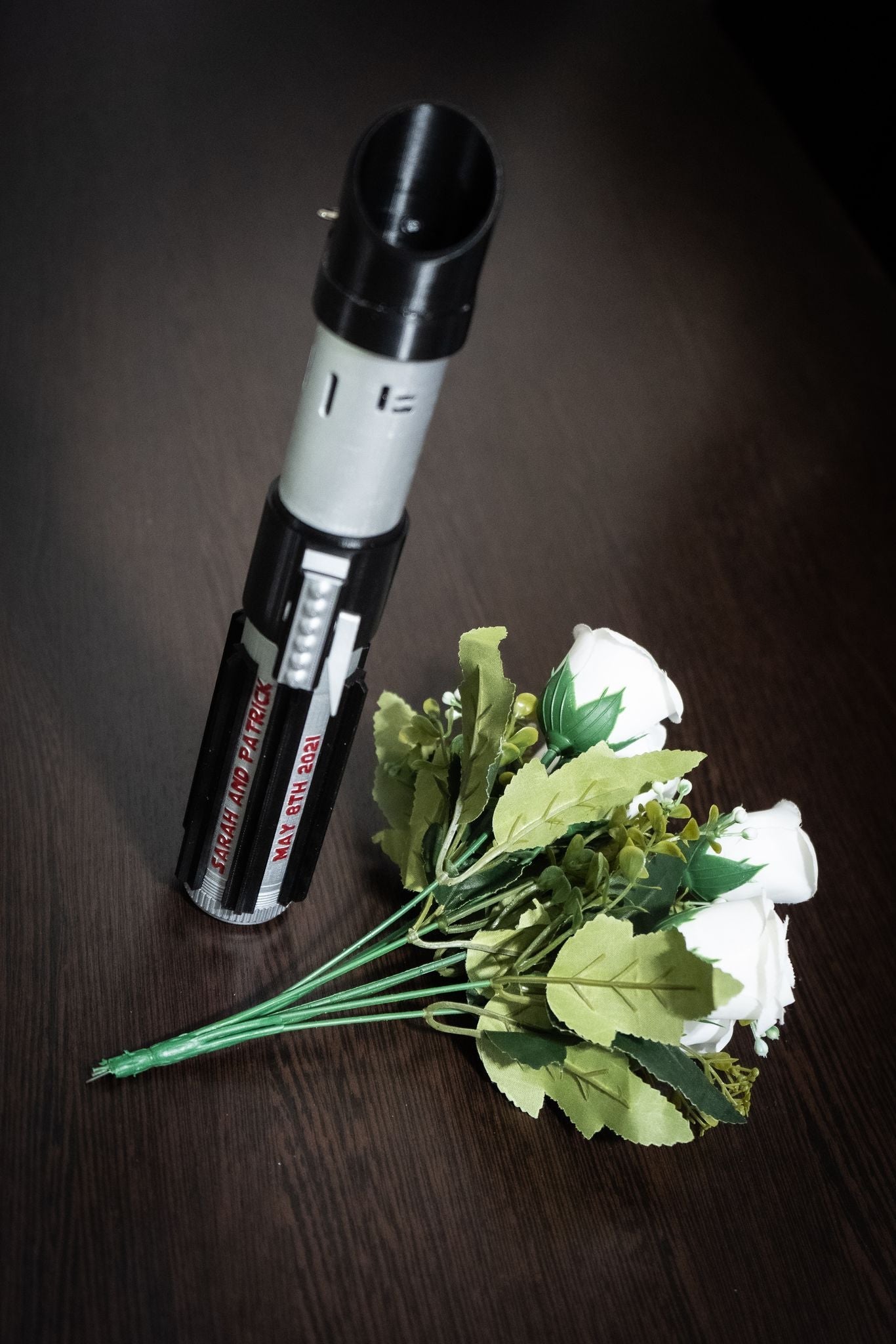 Star Wars inspired Darth Vader lightsaber bridal bouquet holder with white roses and green leaves on dark wooden surface
